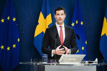 Forssell is talking during a press conference, standing in front of the Swedish flag and the EU flag