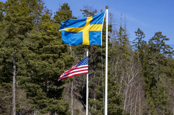 A swedish and an american flag blows in the wind with trees in the background.