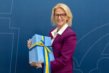 Minister for Finance in a purple suit, holding the budget bill in her hand. She is standing in front of the Government Offices’ backdrop.