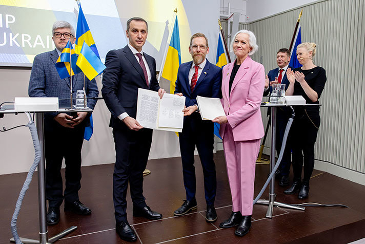 Three ministers, surrounded by people and Swedish and Ukrainian flags, hold up signed agreements.