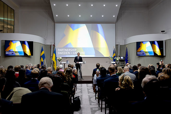 A man gives a speech on stage in a packed conference room. The Swedish and Ukrainian flags, and the text ‘Partnership Sweden-Ukraine’, are shown on the screen behind him.