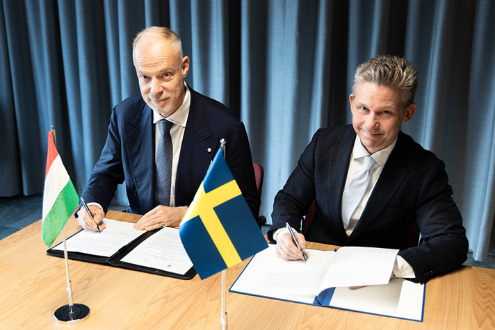 From the left Kristóf Szalay-Bobrovniczky and to the right Pål Jonson sitting at a table signing papers. At the table is the Hungarian flag and the Swedish flag.