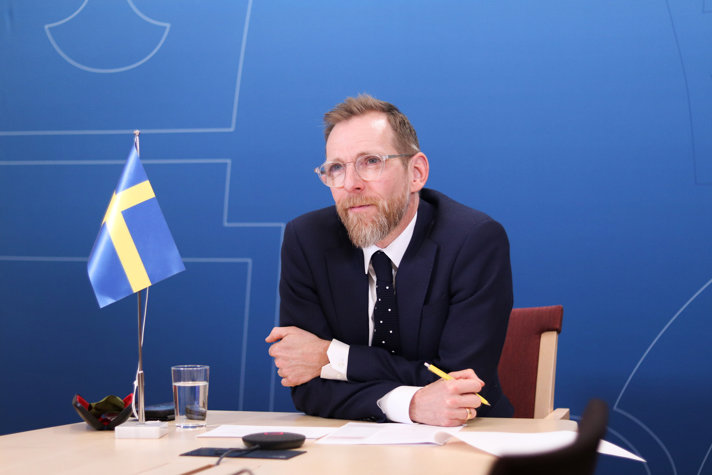 Photo of Jakob Forssmed behind a desk. There is a Swedish flag beside him on the table.